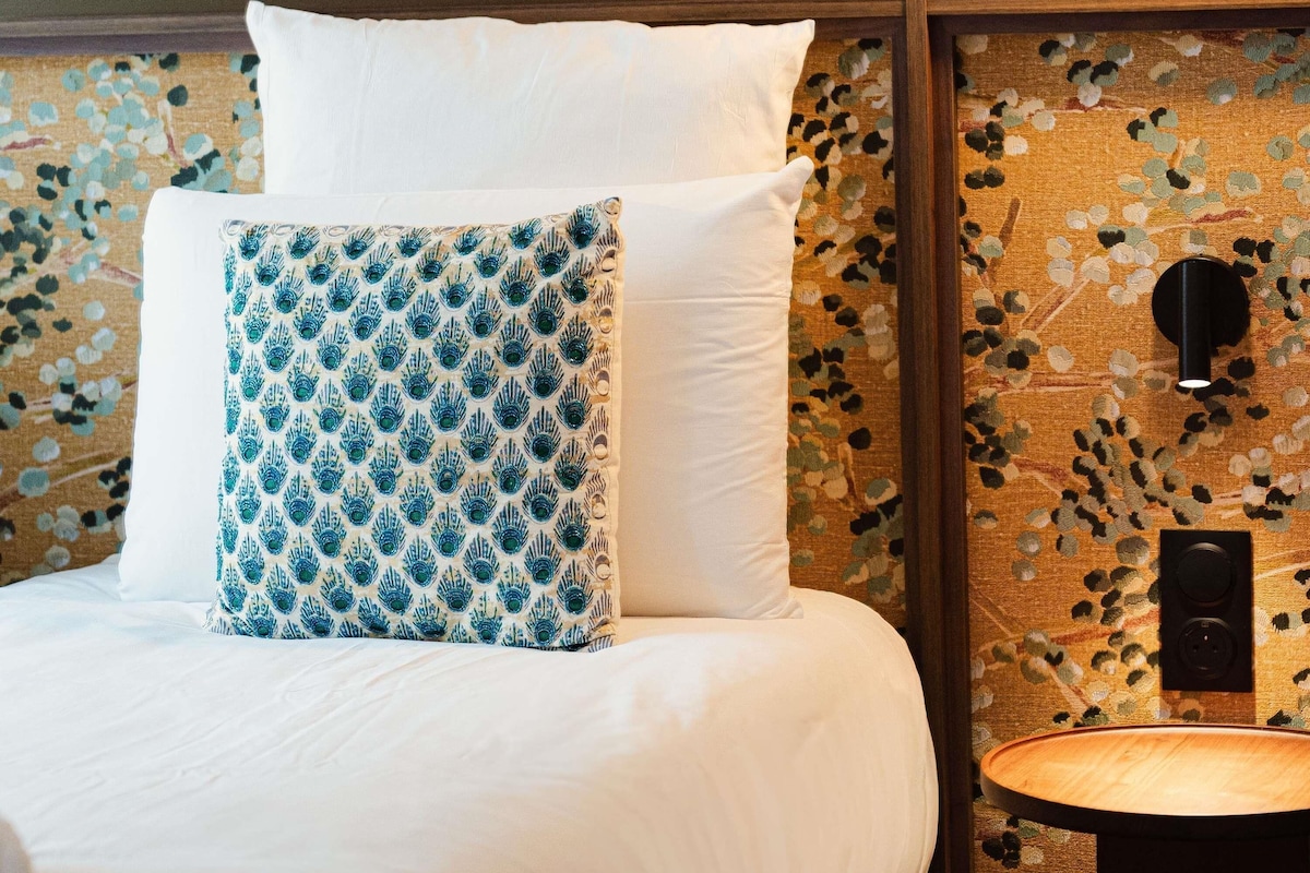 Un lit avec une literie blanche et un coussin à motifs, une petite table de chevet et un mur orné d’un motif floral, ainsi qu’un interrupteur.
