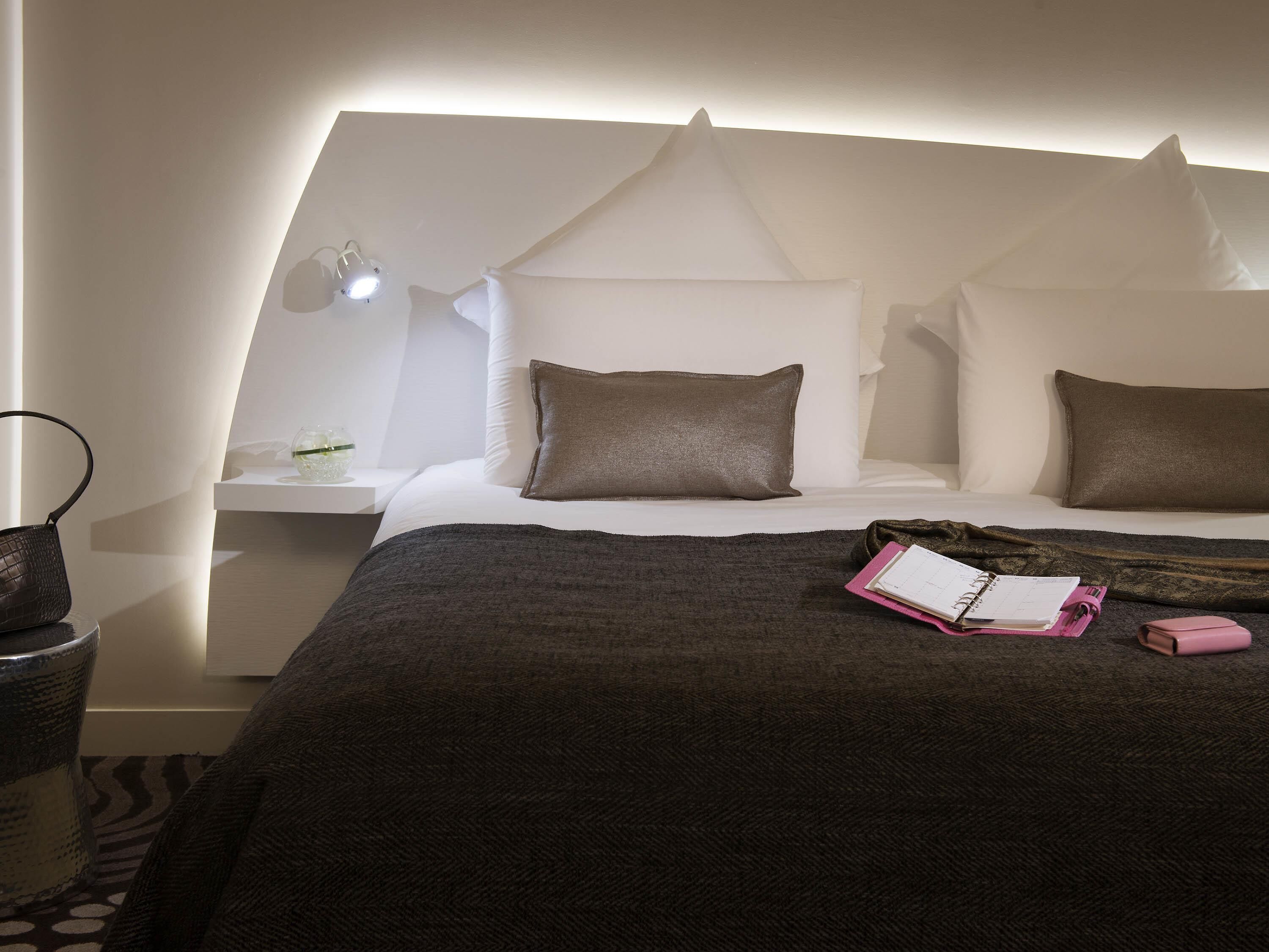 Habitación de hotel ordenada con cama, mesita de noche, lámpara y una pequeña mesa con un recipiente de vidrio.