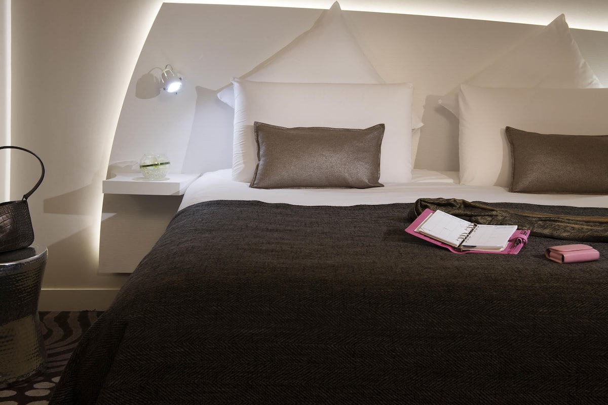 Habitación de hotel ordenada con cama, mesita de noche, lámpara y una pequeña mesa con un recipiente de vidrio.