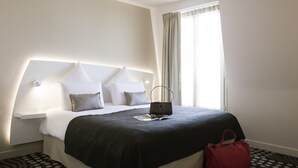 A hotel room with a large bed, a bedside table, a nightstand, a window with curtains, and a red handbag on the floor.