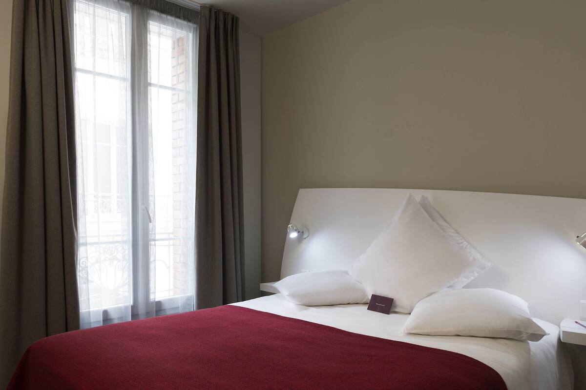 Een hotelkamer met een groot bed, nachtkastje, lamp en raam met gordijnen.