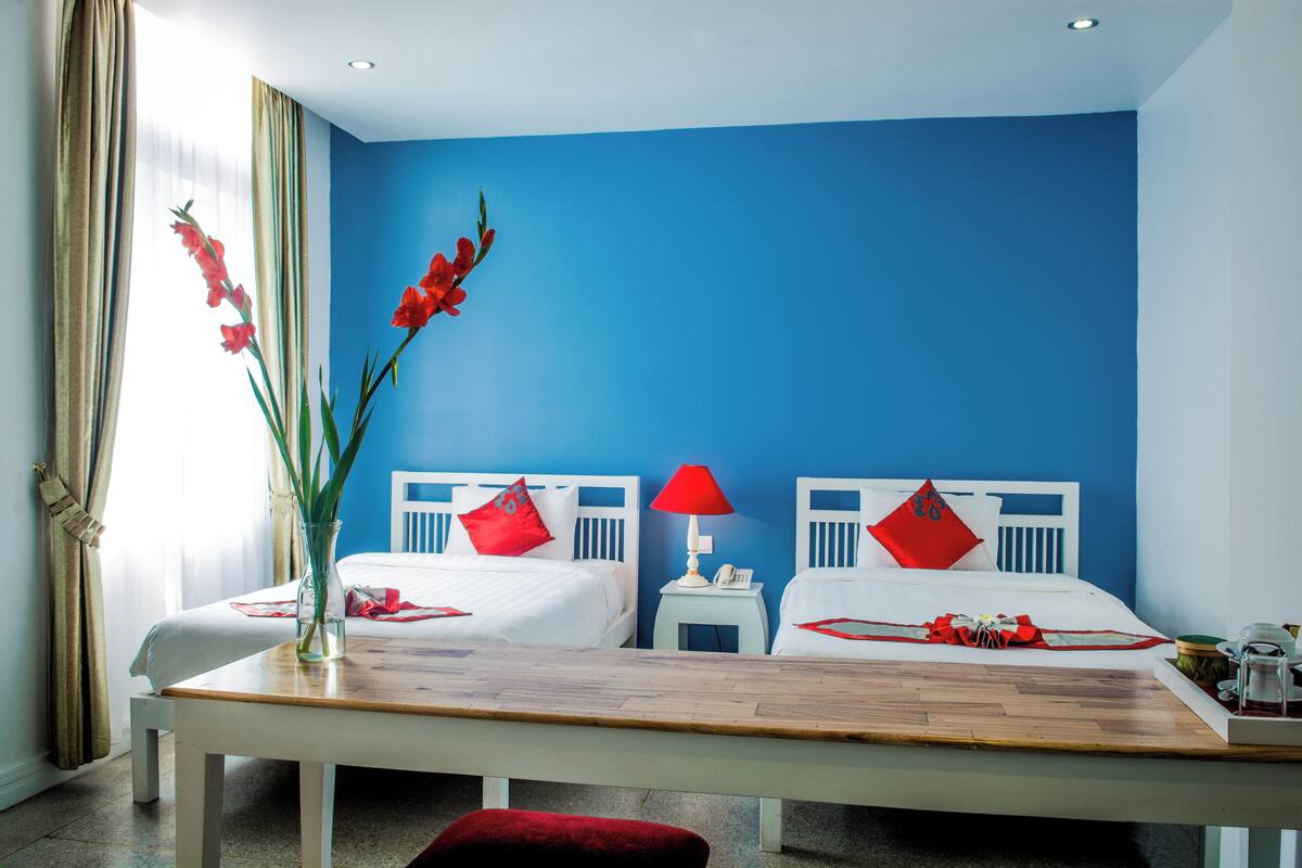 Ein Hotelzimmer mit zwei Betten, einem Holztisch, einer Vase mit roten Blumen und einer roten Lampe.