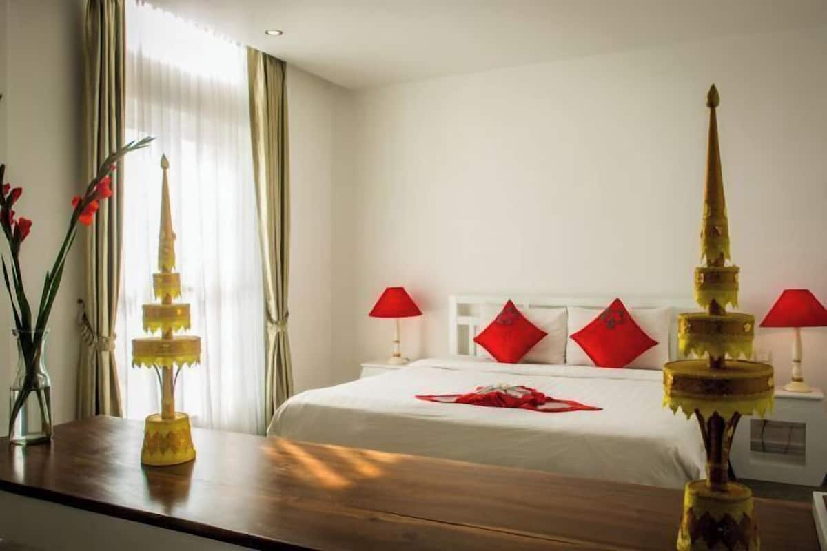 Ein Hotelzimmer mit einem Bett, zwei Nachttischen, einer Lampe, einer Vase mit Blumen und einem Fenster mit Vorhängen.