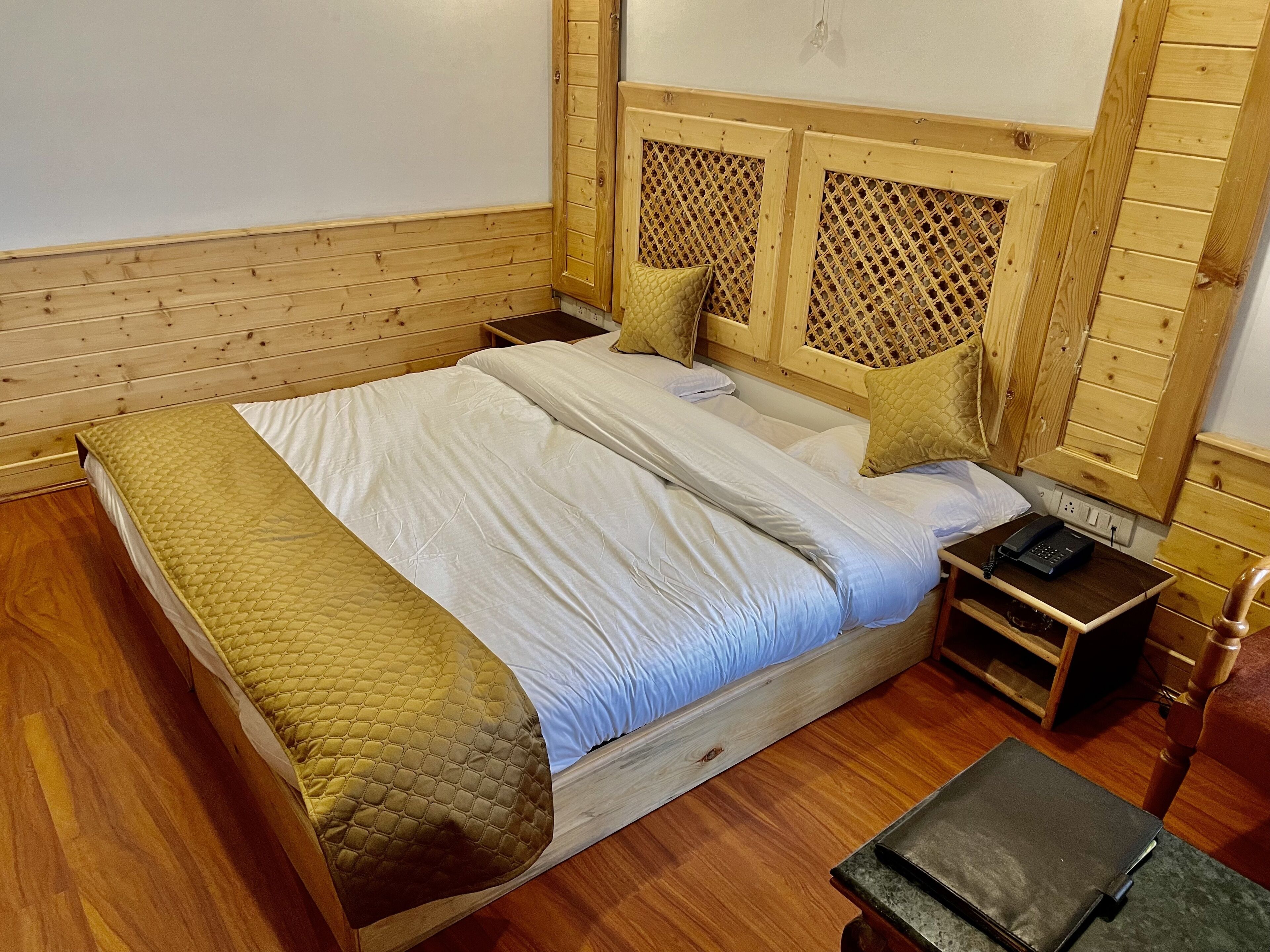 A wooden-framed bed with a quilted bedspread and pillows, a bedside table with a phone, and a small wooden nightstand.