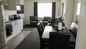 Een moderne keuken met witte kasten, een ingebouwde oven en een koffiezetapparaat. Er is een eethoek met een zwarte tafel en stoelen, en een vaas met bloemen op de tafel.