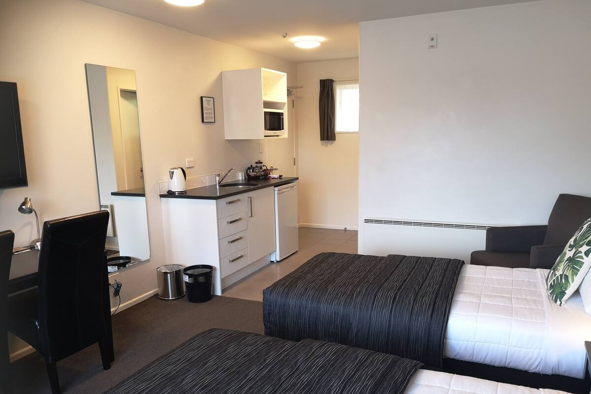 A hotel room with two beds, a kitchenette, a desk, and a TV.