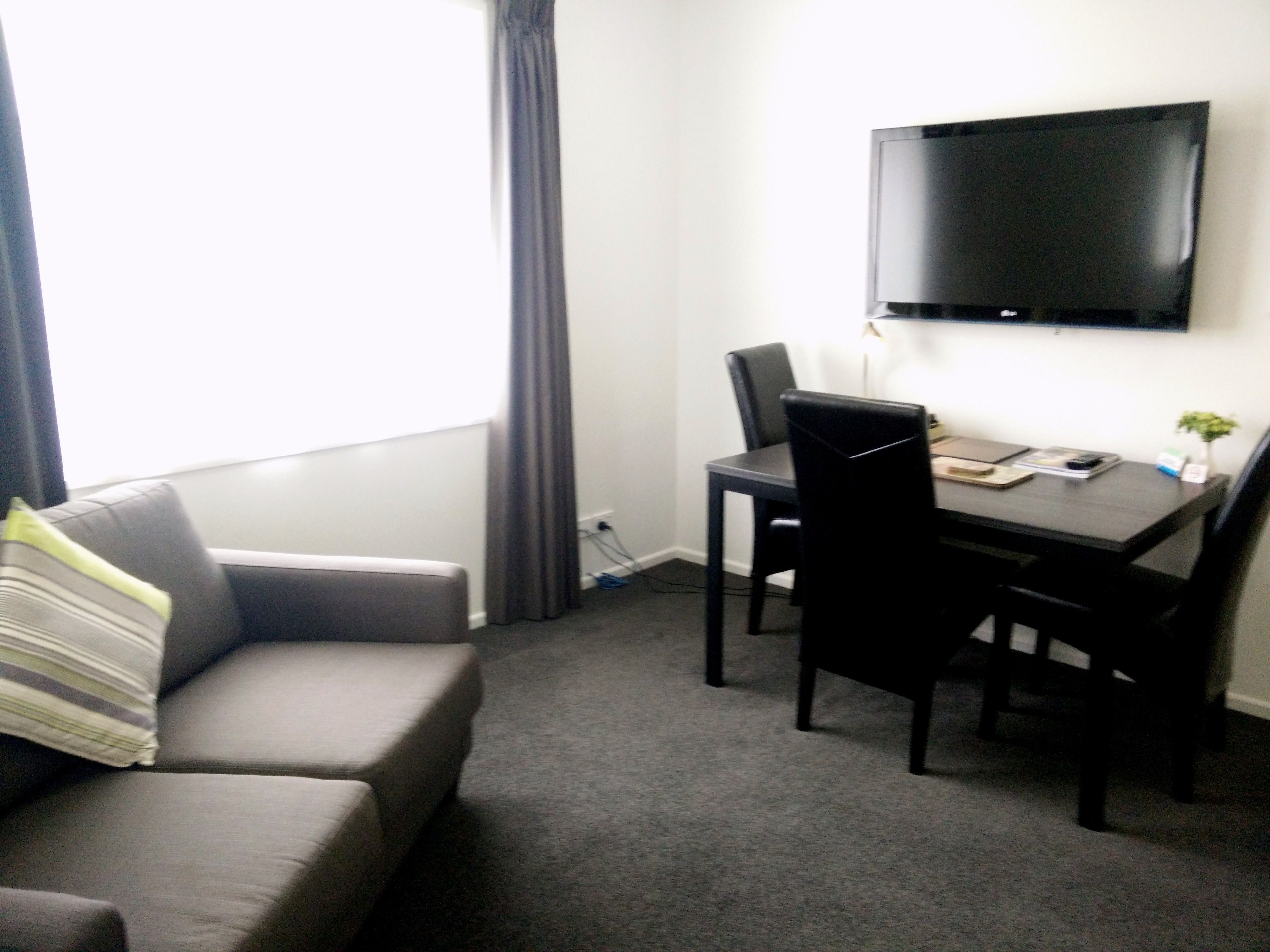 A small room with a grey sofa, a dining table with two chairs, a flat-screen TV mounted on the wall, and a window with curtains.