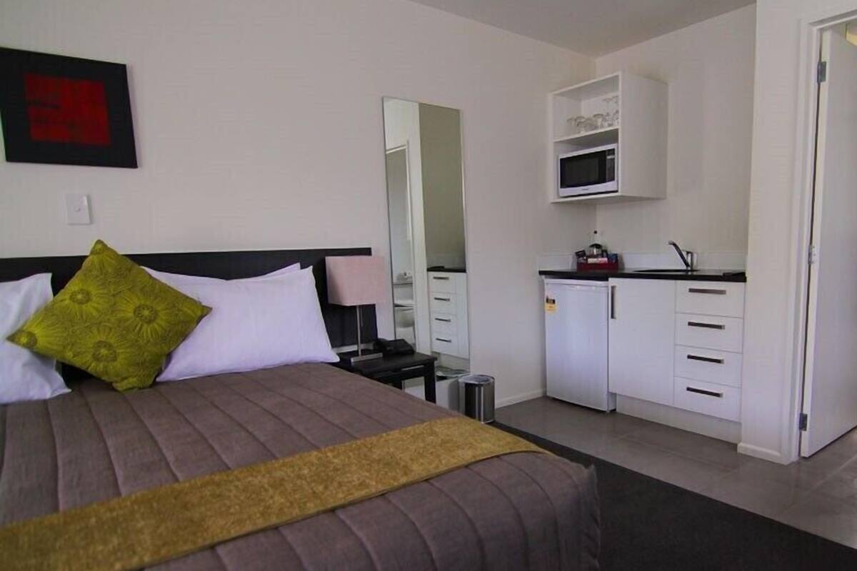 A hotel room with a bed, a bedside table, a lamp, a small kitchenette, and a mirror.