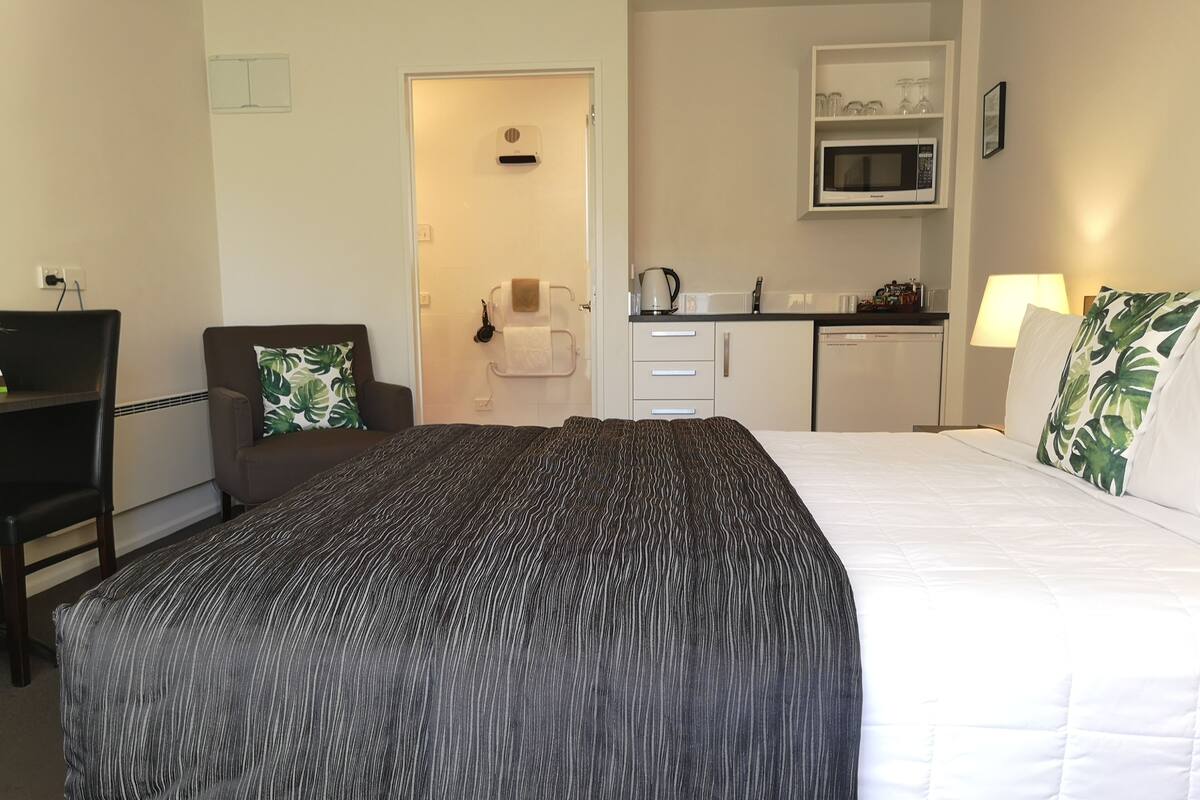 A hotel room with a bed, a desk, a chair, a microwave, and a small kitchenette.