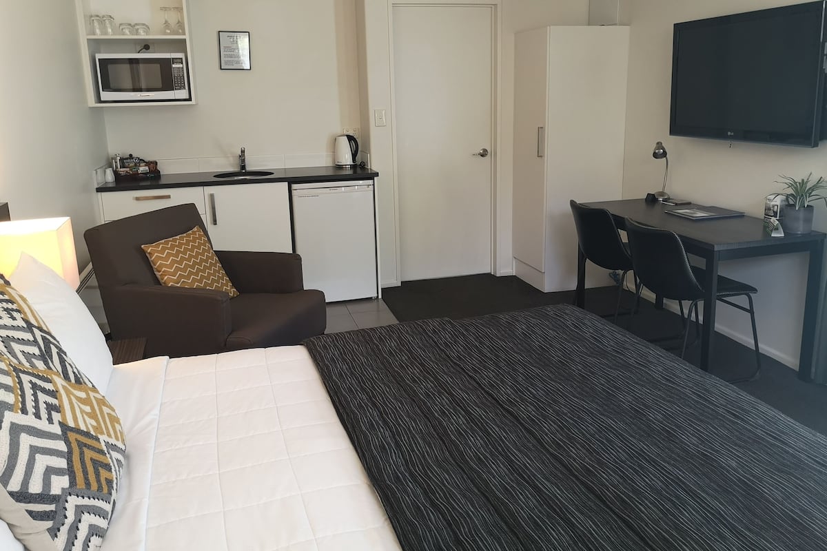 A hotel room with a bed, a desk, a chair, a kitchenette, and a TV.
