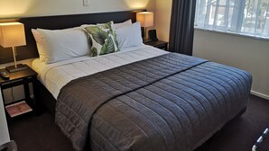 Een net opgemaakt bed met een donkere deken, twee bedlampen, een afstandsbediening en een aan de muur bevestigd kunstwerk.