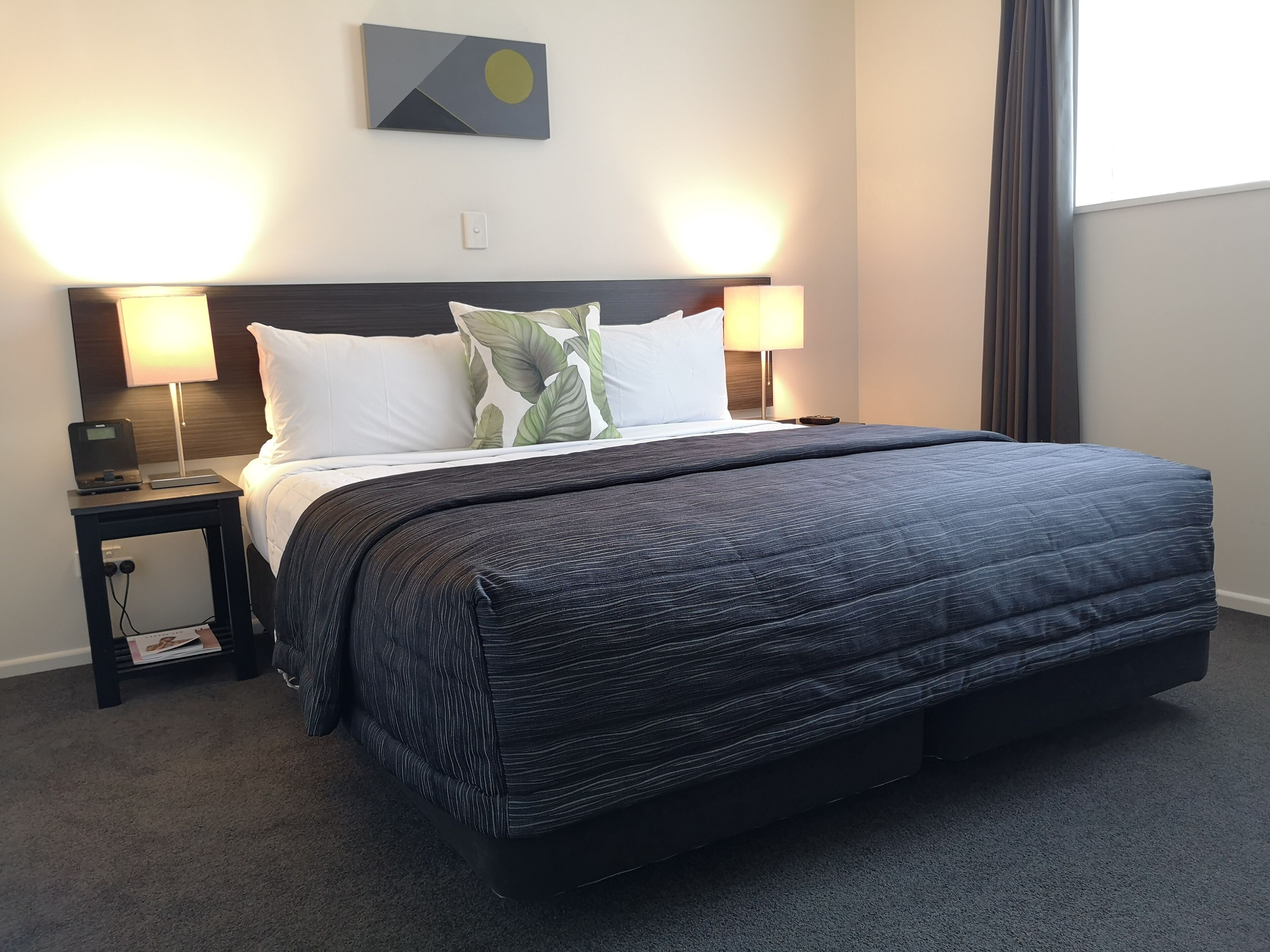 A hotel room with a large bed, two bedside lamps, a nightstand, a wall-mounted artwork, and a window with curtains.