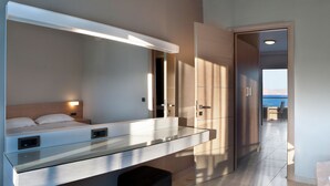 Habitación de hotel moderna con una cama grande, una mesita de noche de cristal, televisión integrada y vista al mar a través de una puerta abierta.