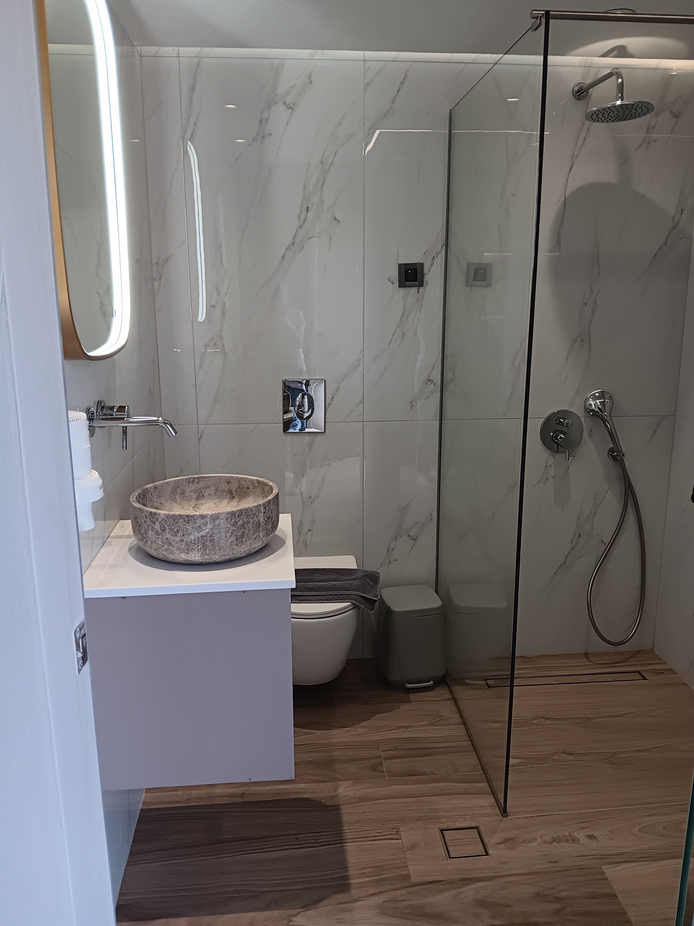Ein modernes Badezimmer mit Marmorwand, einer Duschkabine aus Glas, einem weißen Waschbecken mit Steintonne und einer Toilette.