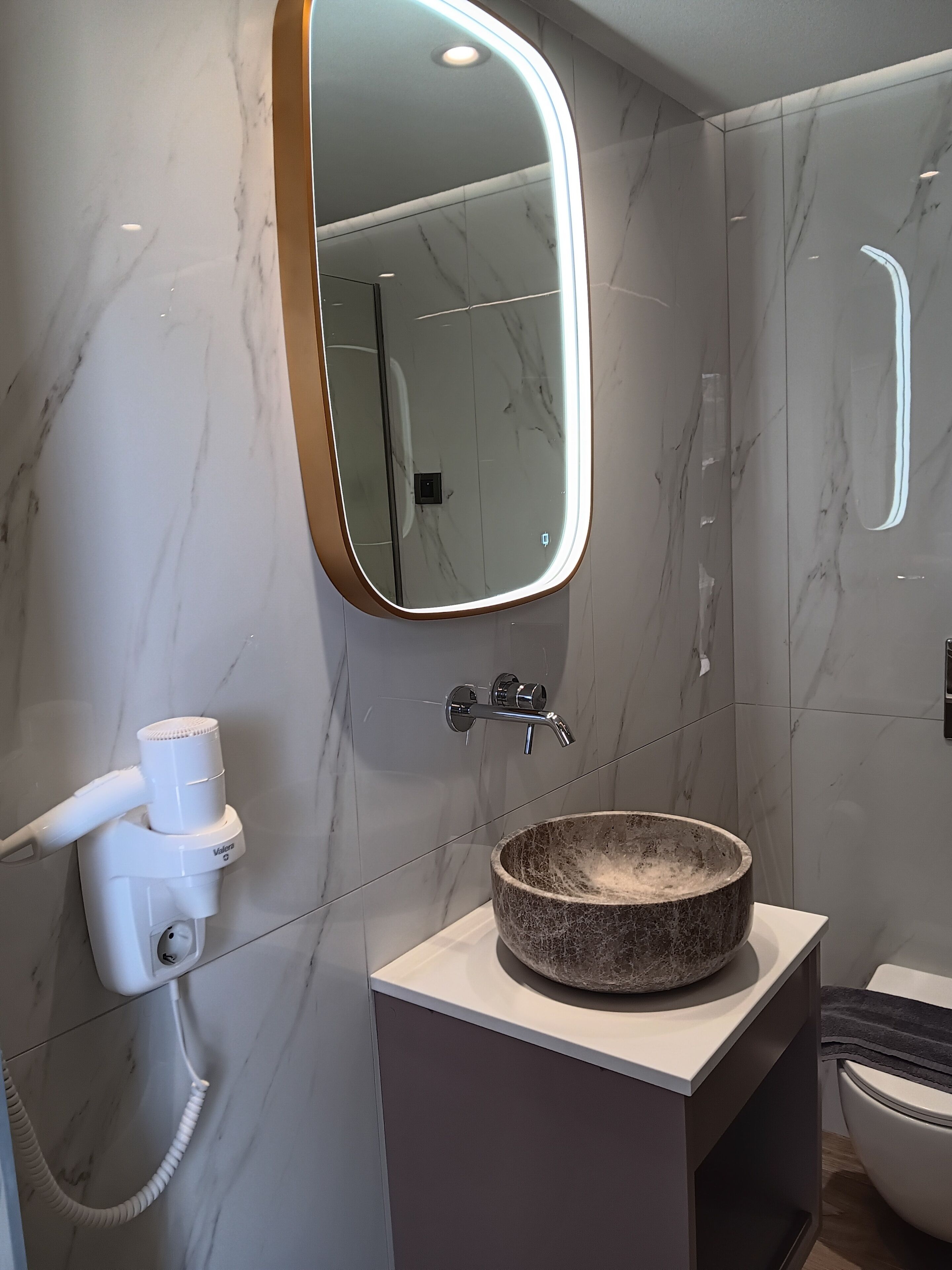A modern bathroom with a marble wall, a round basin, a handheld hair dryer, and a mirror with a light strip.