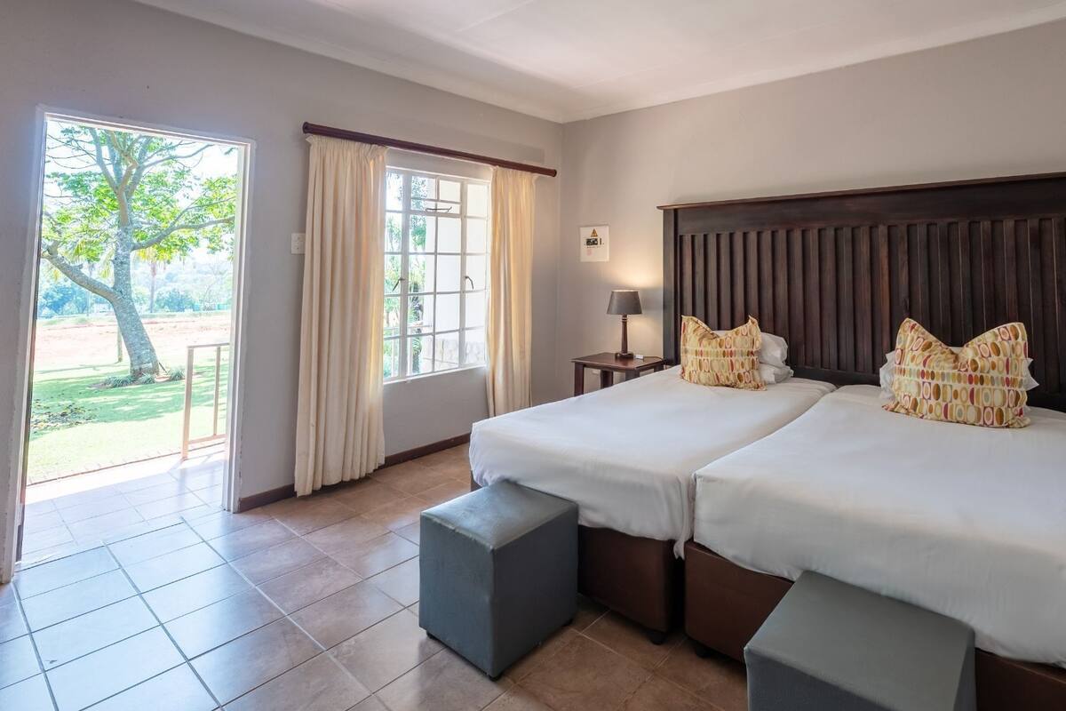 A hotel room with two beds, a window with curtains, a nightstand, and a view of greenery outside.