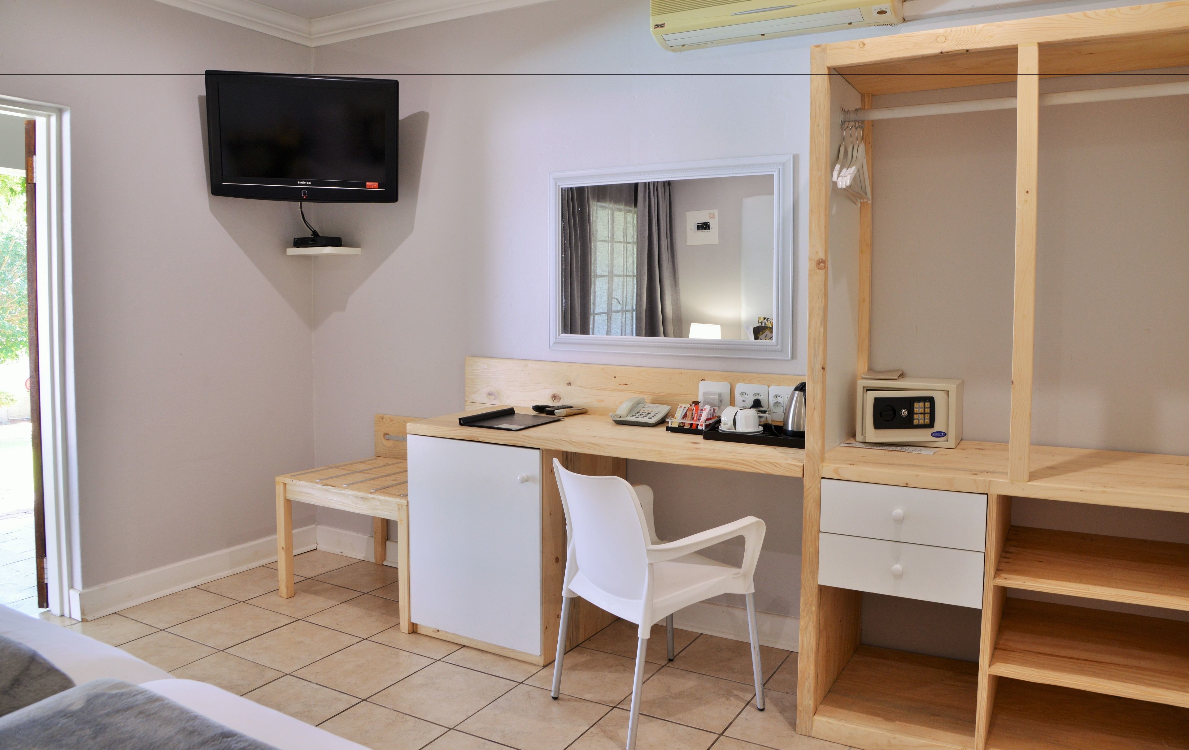 A bedroom with a wooden desk, a white chair, a TV mounted on the wall, a mirror, and a wardrobe.