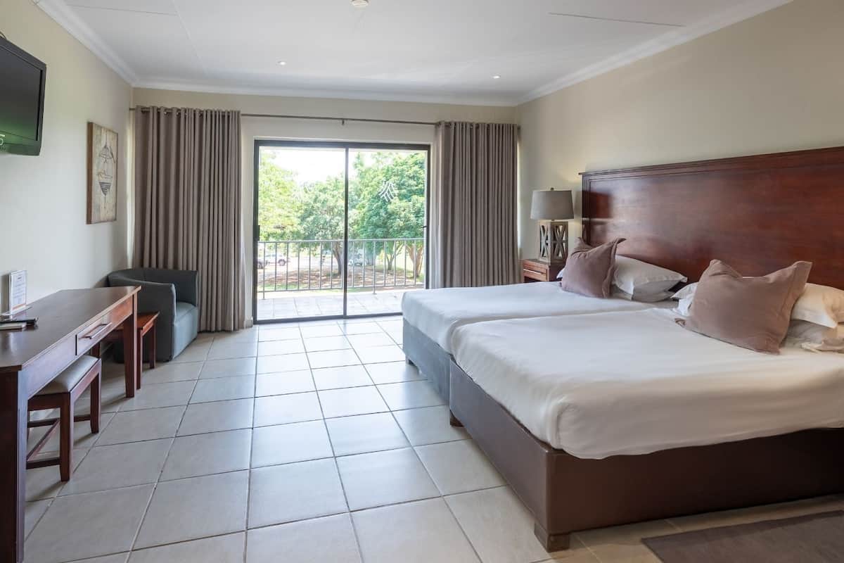 A hotel room with a large bed, a desk, and a chair. There is a balcony with curtains and a view of trees.