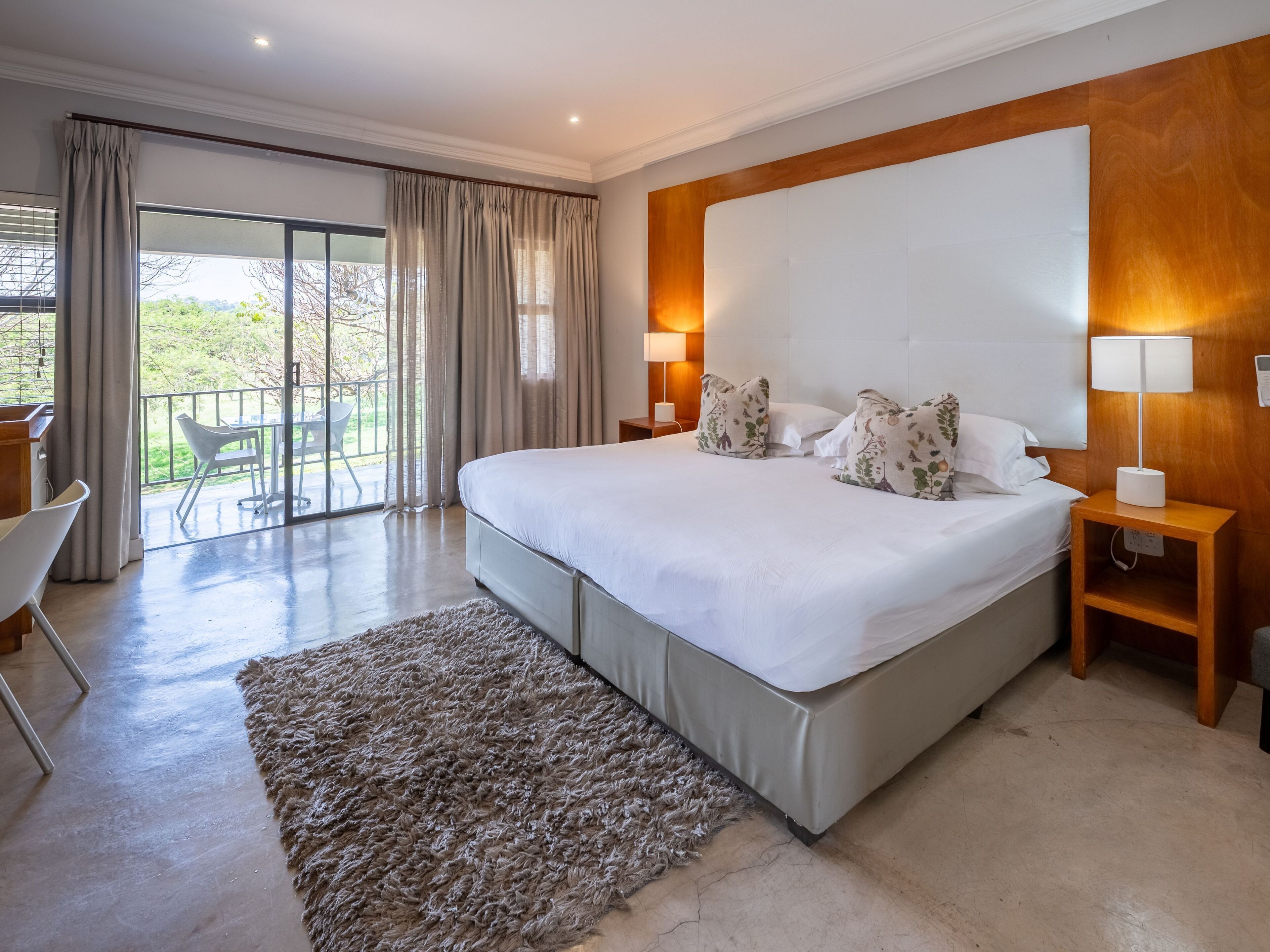 A spacious bedroom with a large bed, two bedside lamps, a wooden headboard, and a view of a balcony.