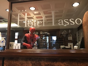 Een hotel receptie met een grote spiegel die een Spider-Man figuur weerspiegelt. De receptiebalie is van hout met een glazen blad. De hotelnaam 'Hotel Tasso' is zichtbaar boven de spiegel.