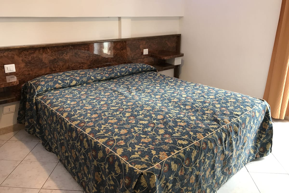 A room with a bed, a wooden headboard, a nightstand, and a wall-mounted light switch.