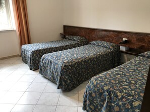 Een kamer met drie bedden, elk met een bloemenprint sprei. Er is een houten hoofdbord, een klein tafeltje met een telefoon en een doos, en een raam met gordijnen.