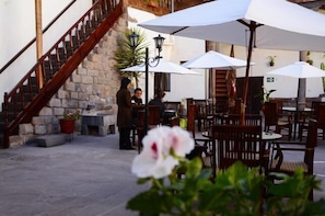 Un área de café al aire libre con paredes de piedra, mobiliario de madera y sombrillas blancas.