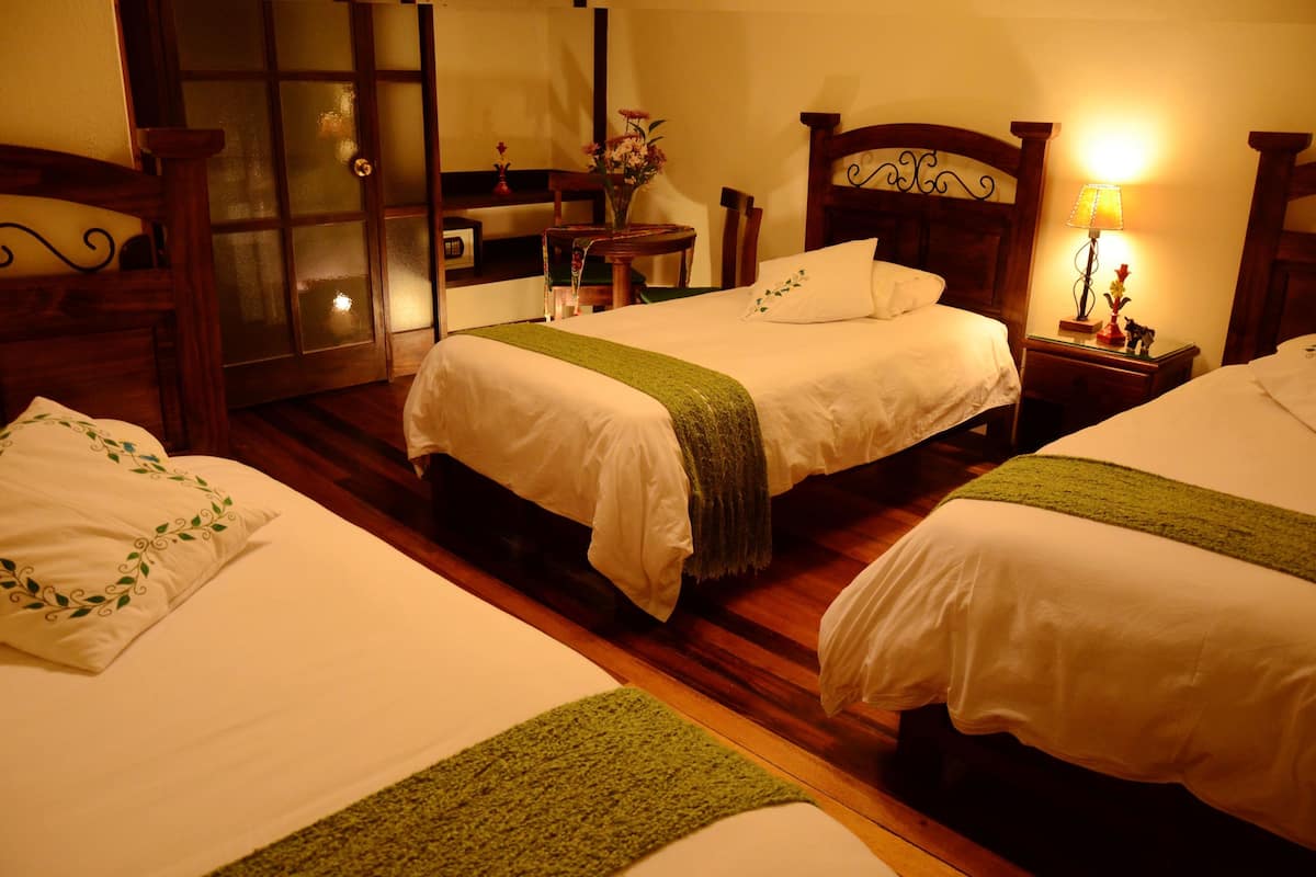 A hotel room with two beds, wooden headboards, a small table with a vase of flowers, and a nightstand with a lamp.