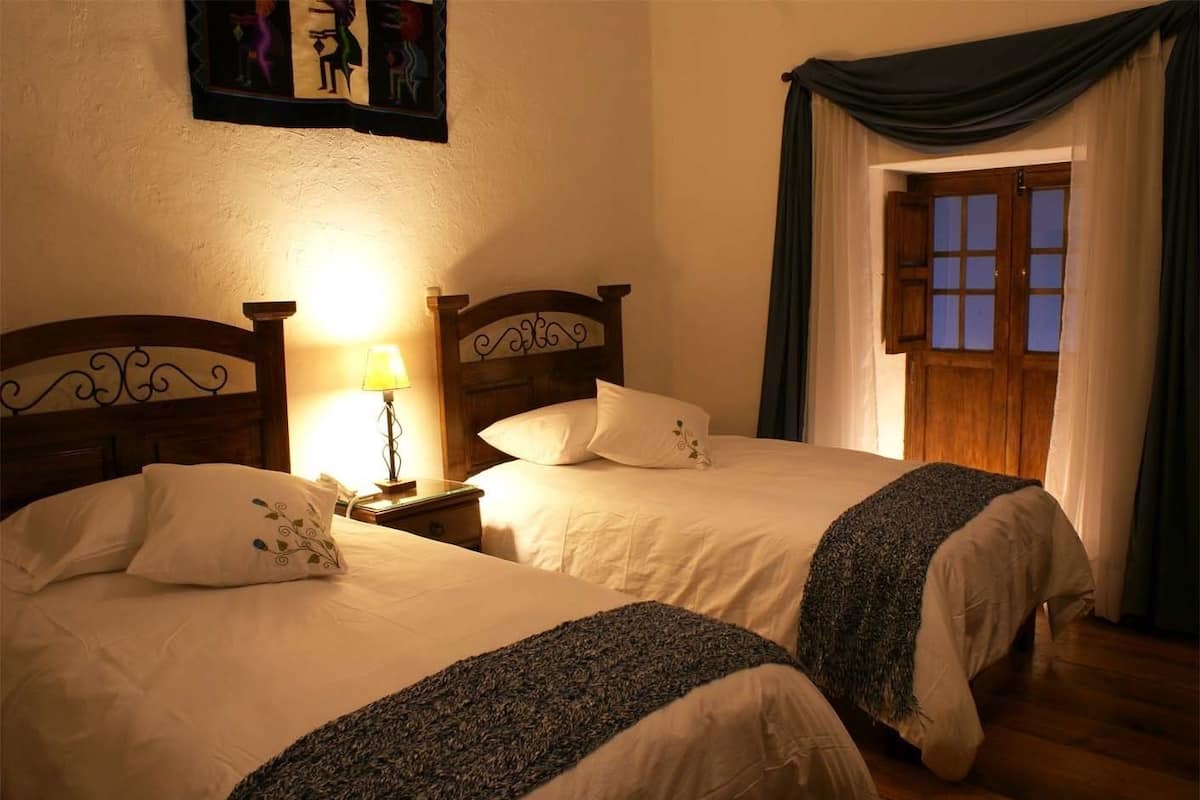 A hotel room with two beds, a nightstand with a lamp, a window with curtains, and a painting on the wall.