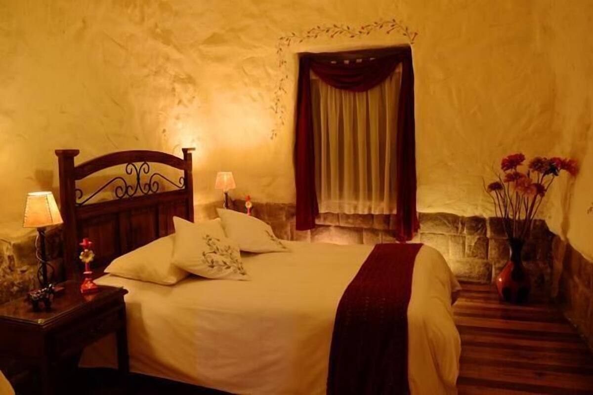 A bedroom with a wooden headboard, a bed with white linens, a nightstand with a lamp, a window with curtains, and a vase with flowers on the floor.