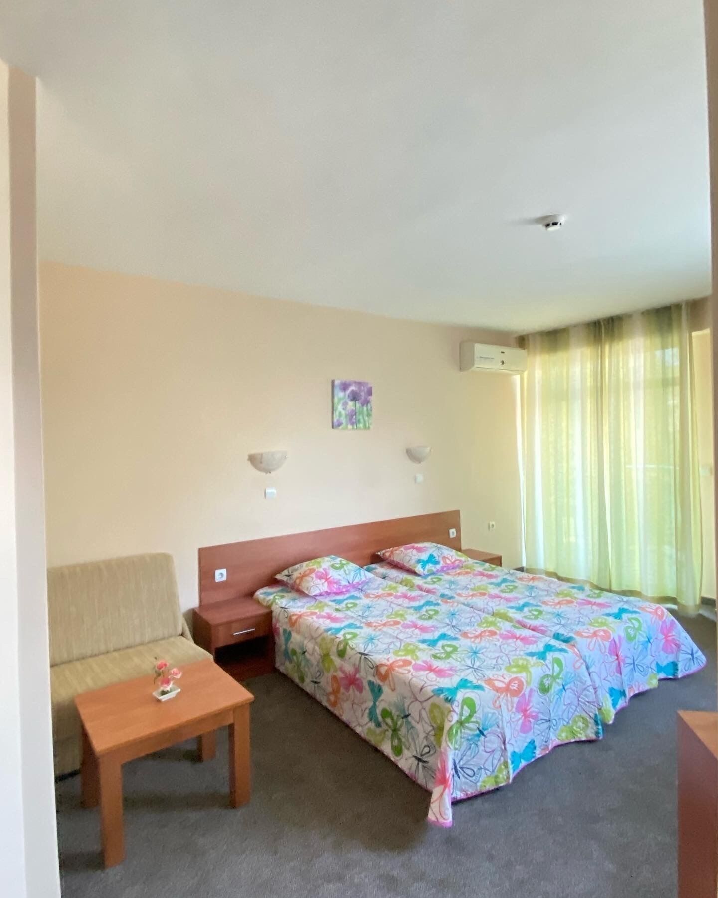 A hotel room with a bed, a sofa, a small table, and a wall-mounted air conditioner.