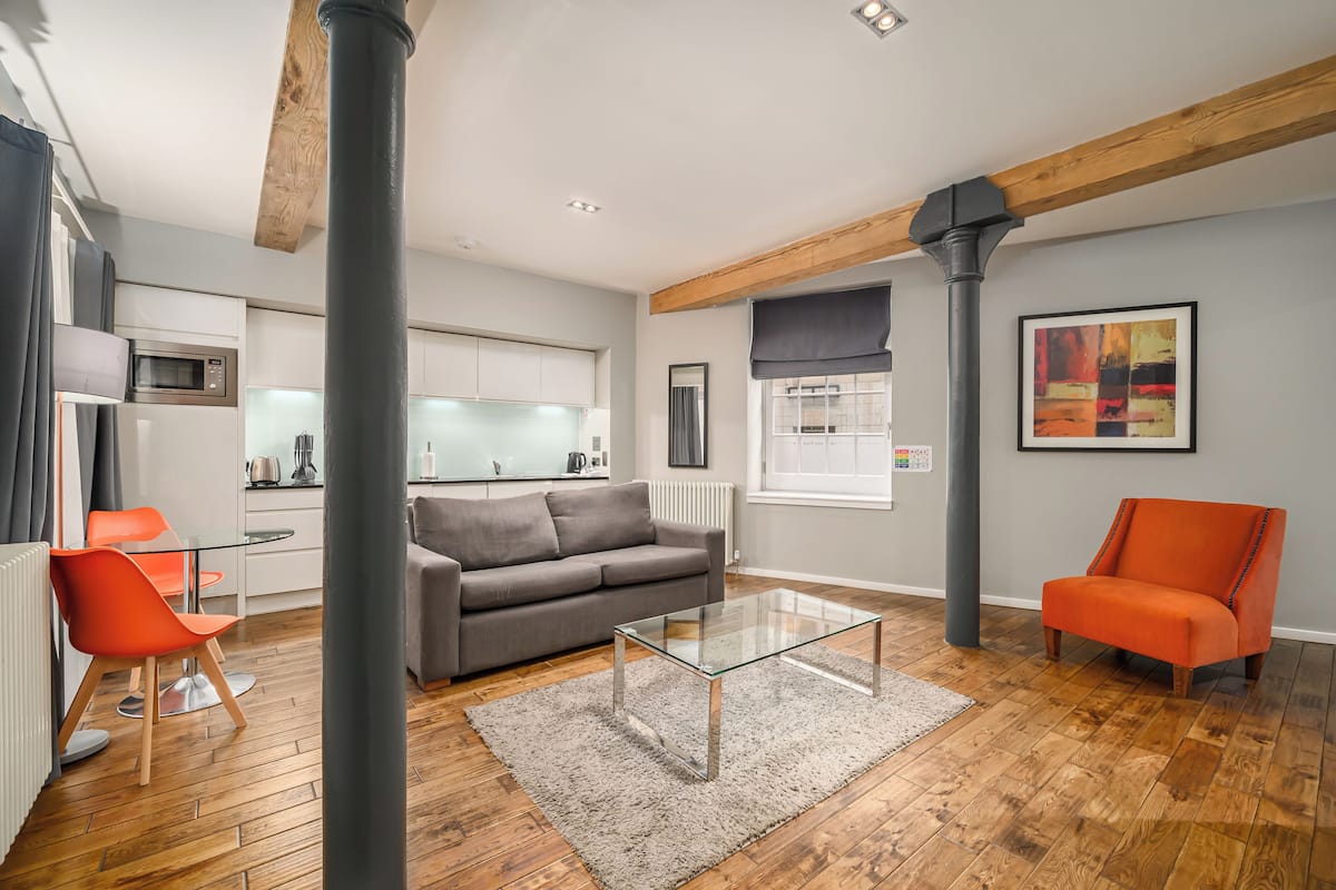 A modern living room with a grey sofa, orange chairs, a glass coffee table, and a kitchen in the background.