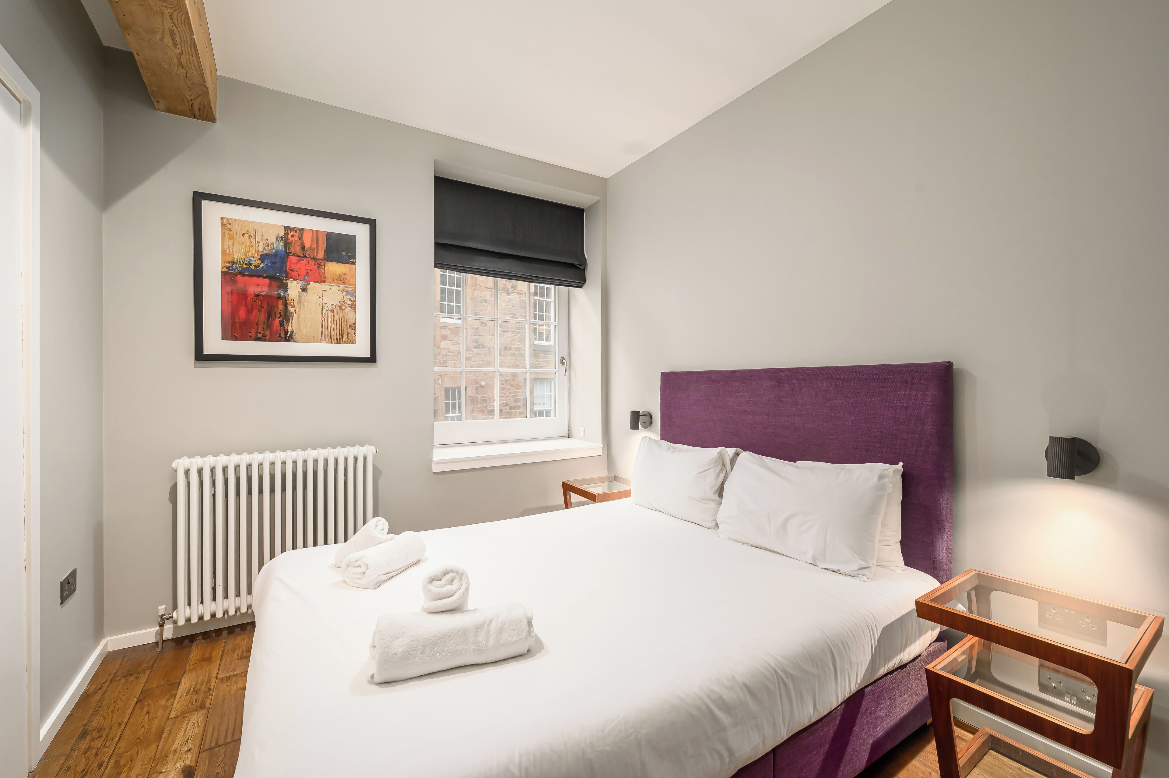 Een net opgemaakt bed met witte lakens en handdoeken, een paars hoofdbord, een nachtkastje met een lamp, een radiator, en een raam met uitzicht op gebouwen.