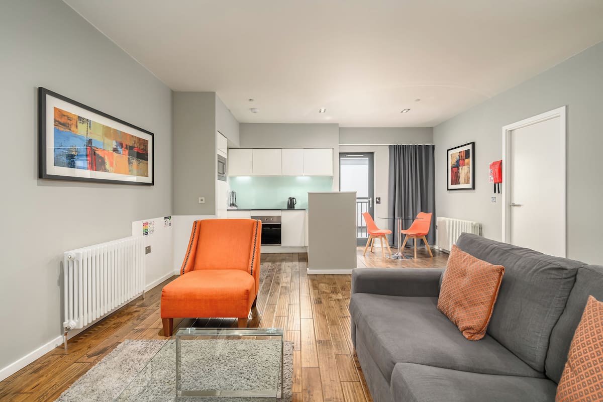 A modern living room with a grey sofa and orange armchair, a kitchen in the background, and a framed abstract painting on the wall.