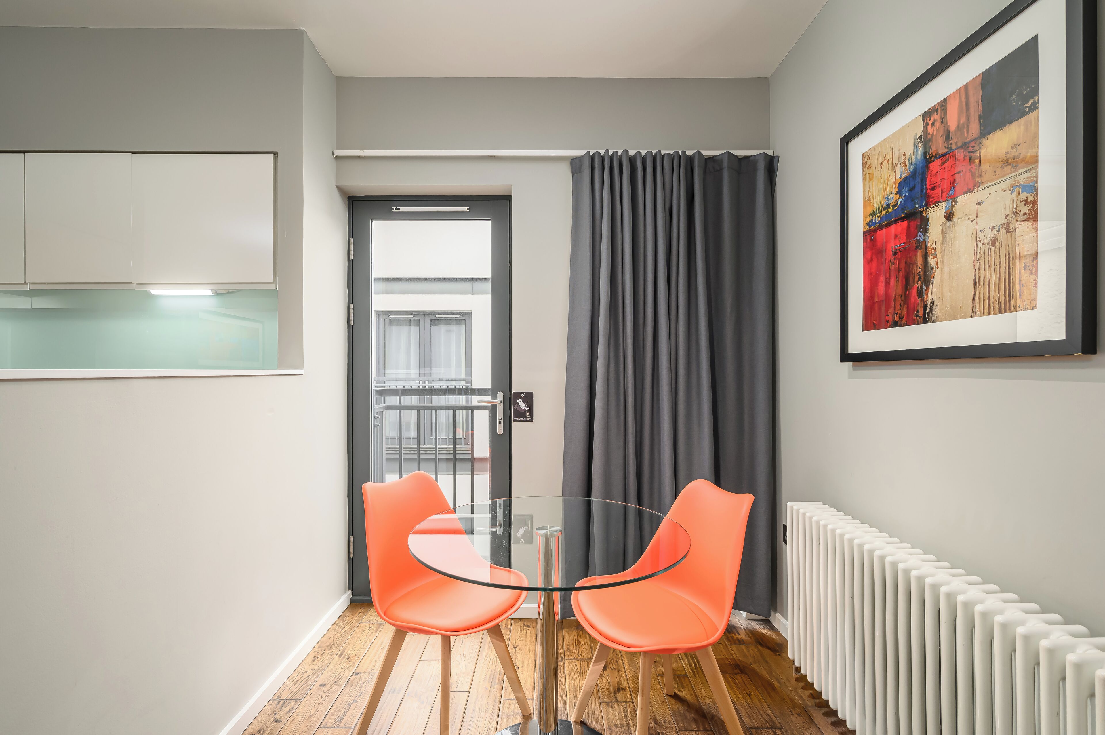 Habitación moderna con una mesa de vidrio, dos sillas color naranja, un radiador y una puerta con vistas.