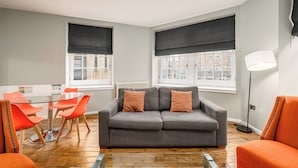 A modern living room with a grey sofa, orange chairs, a glass table, and large windows with black blinds. - Destiny Scotland - The Malt House Apartments (Edinburgh)