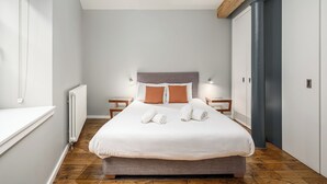 A modern bedroom with a large bed, two nightstands, a radiator, and a wooden ceiling beam. - Destiny Scotland - The Malt House Apartments (Edinburgh)