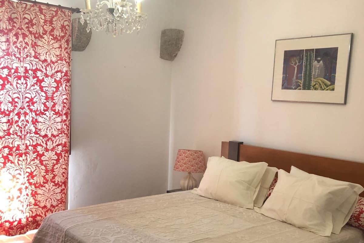 A bedroom with a bed, a chandelier, a red and white patterned curtain, a lamp, and a framed picture on the wall.