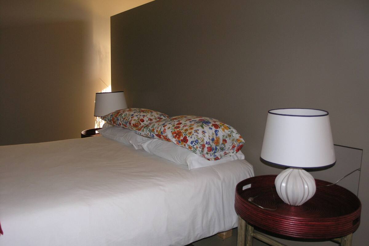 A hotel room with a bed, two pillows, a bedside lamp, and a small table with a lamp.