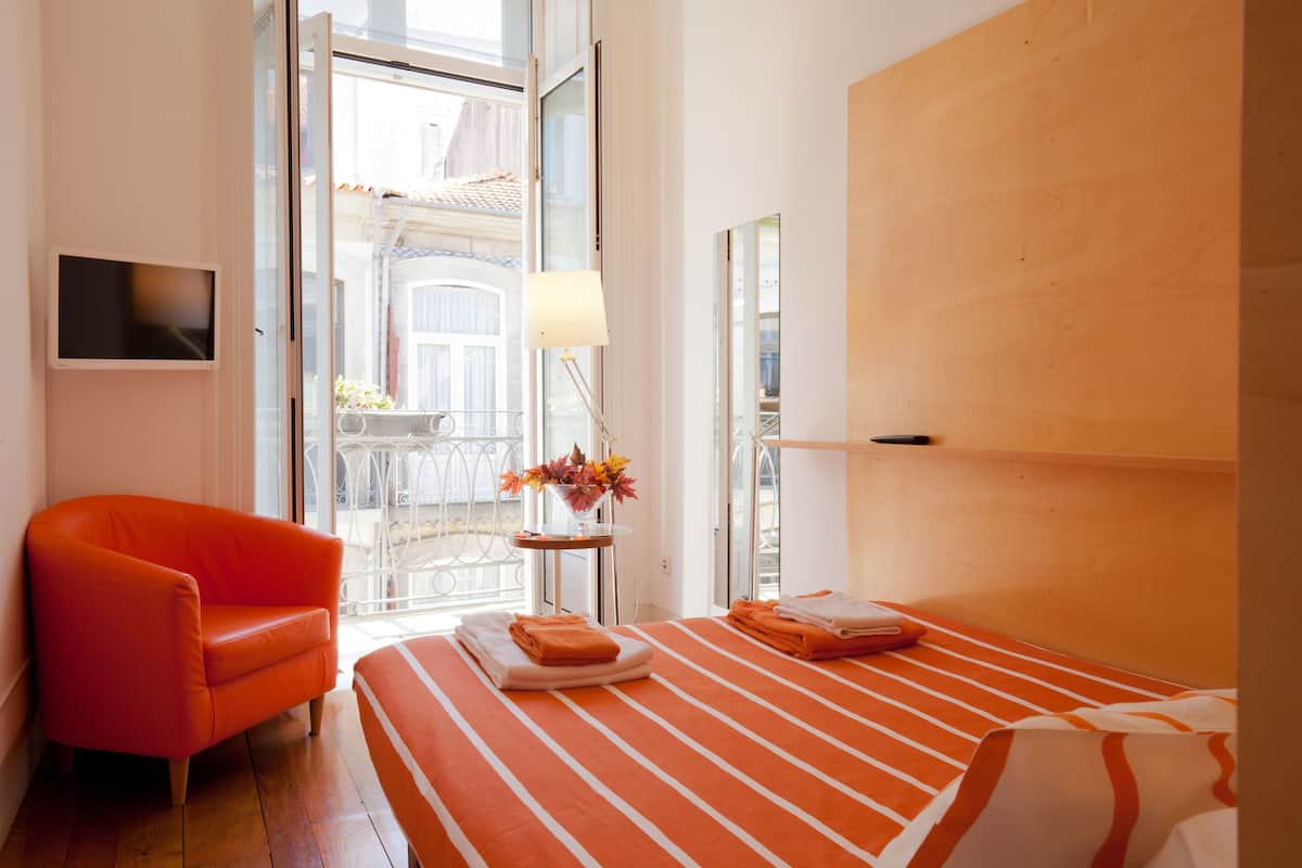A hotel room with a bed, an orange armchair, a small table with a lamp, and a view of the outside.