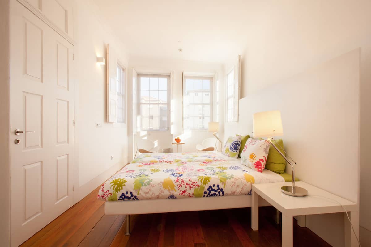 A bedroom with a bed, bedside table, lamp, and a view of the outside through a window.