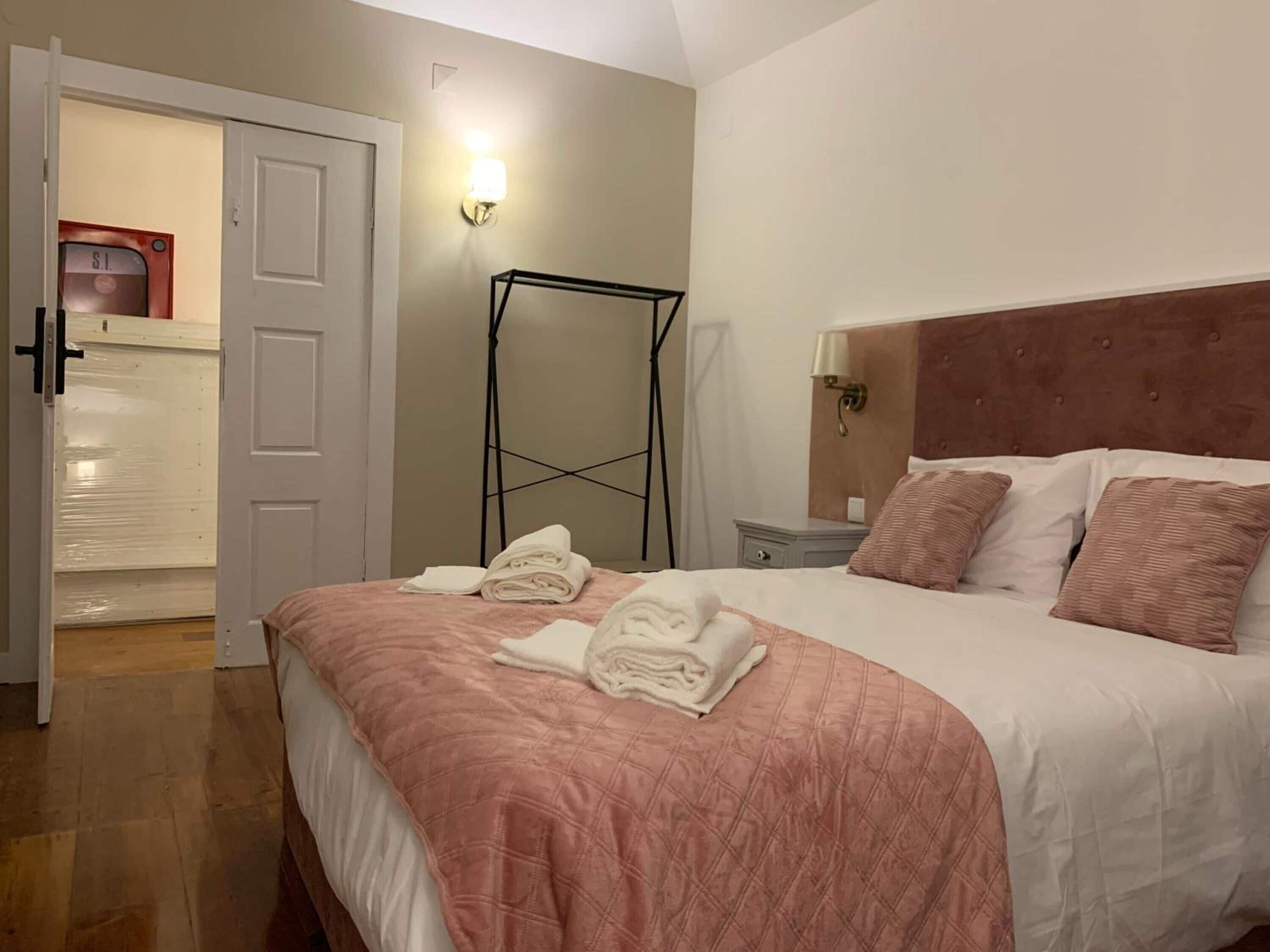 A neatly made bed with a pink quilt, a white comforter, and rolled towels on the bed. There is a wooden floor, a white door, and a wall-mounted light fixture.
