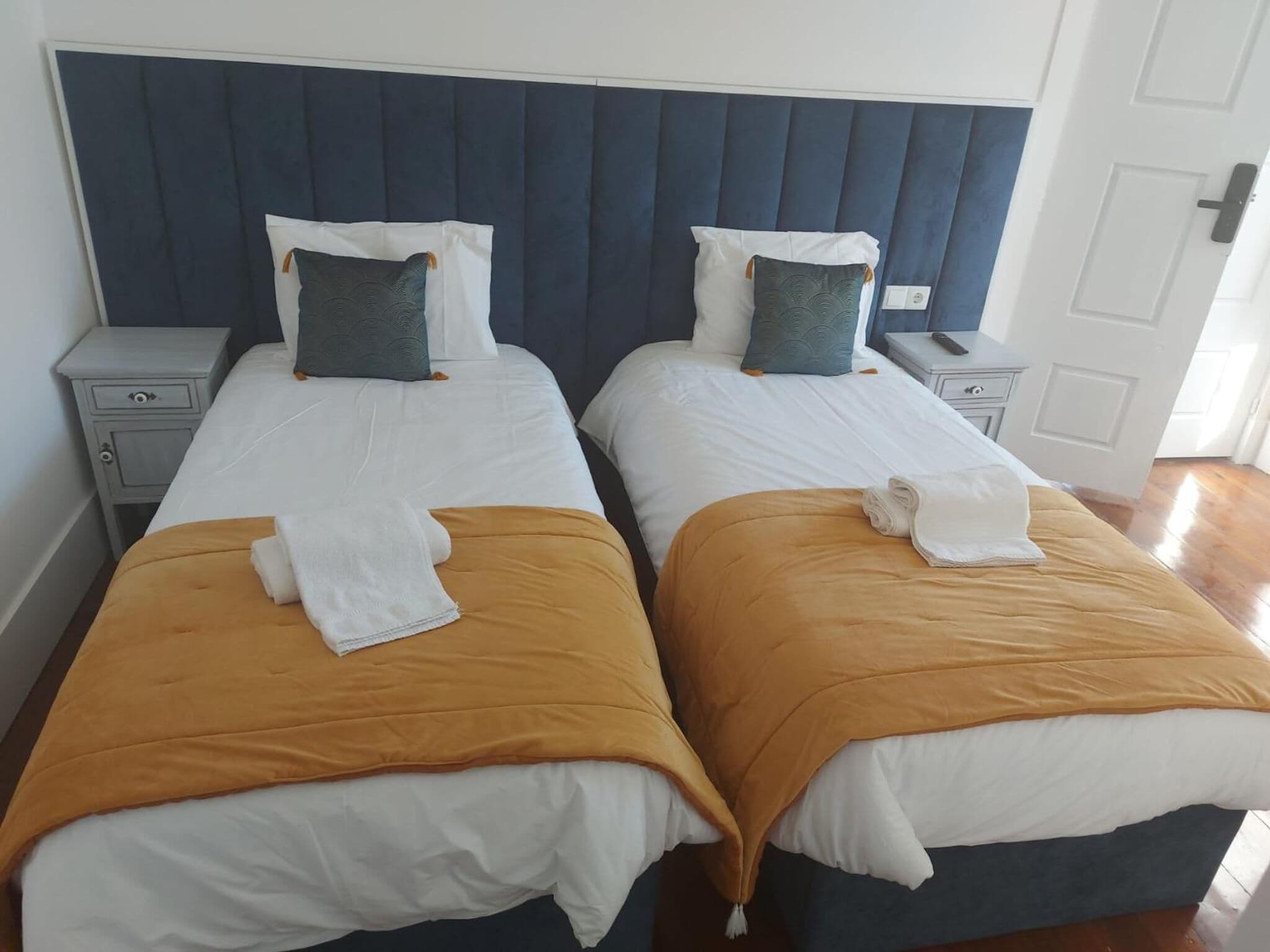 A double room with two beds, each with a white duvet and a mustard-colored blanket. There are two nightstands with drawers and a small table between the beds.