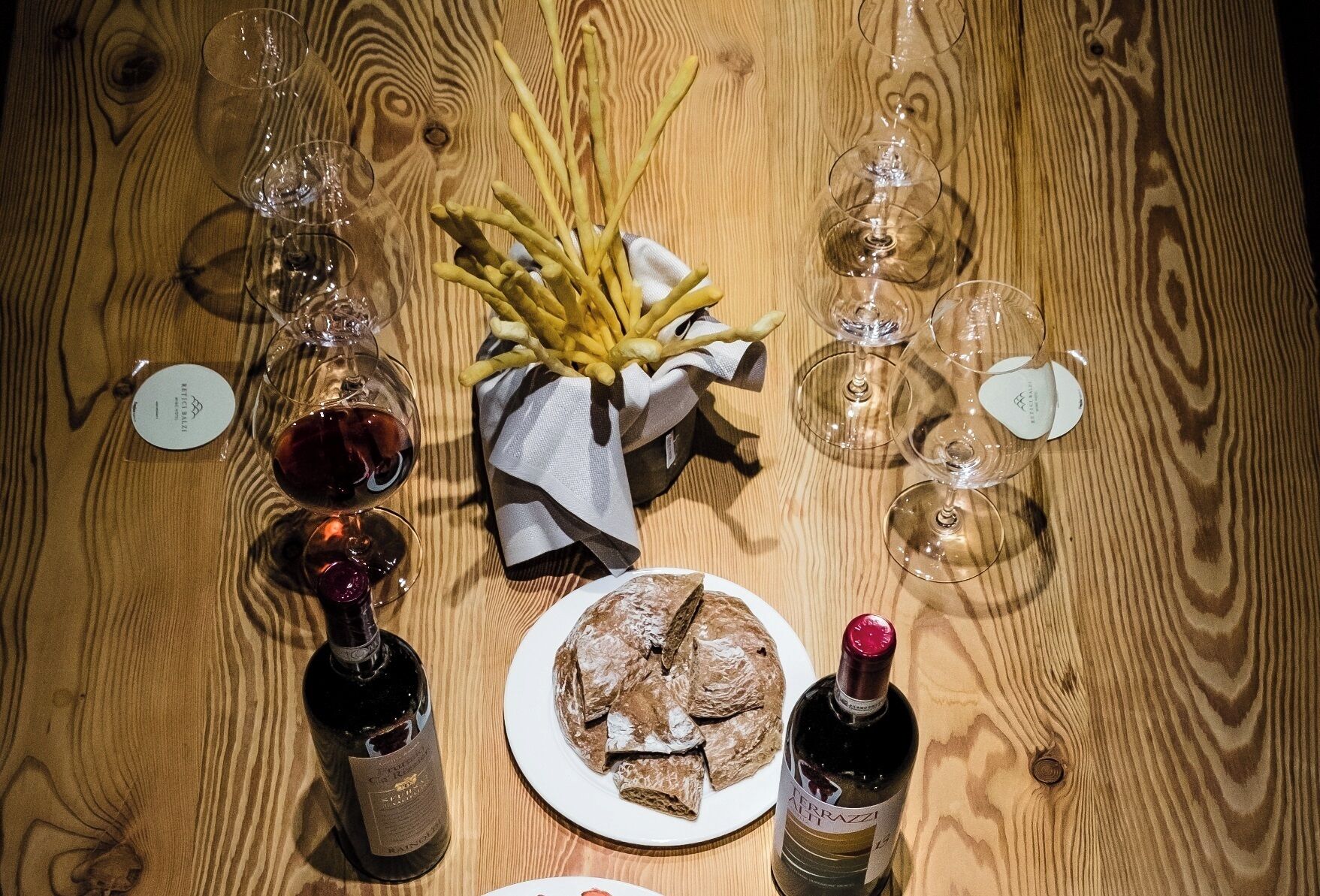 Una mesa de madera con copas de vino, una botella de vino, un plato de pan y una cesta de papas fritas.