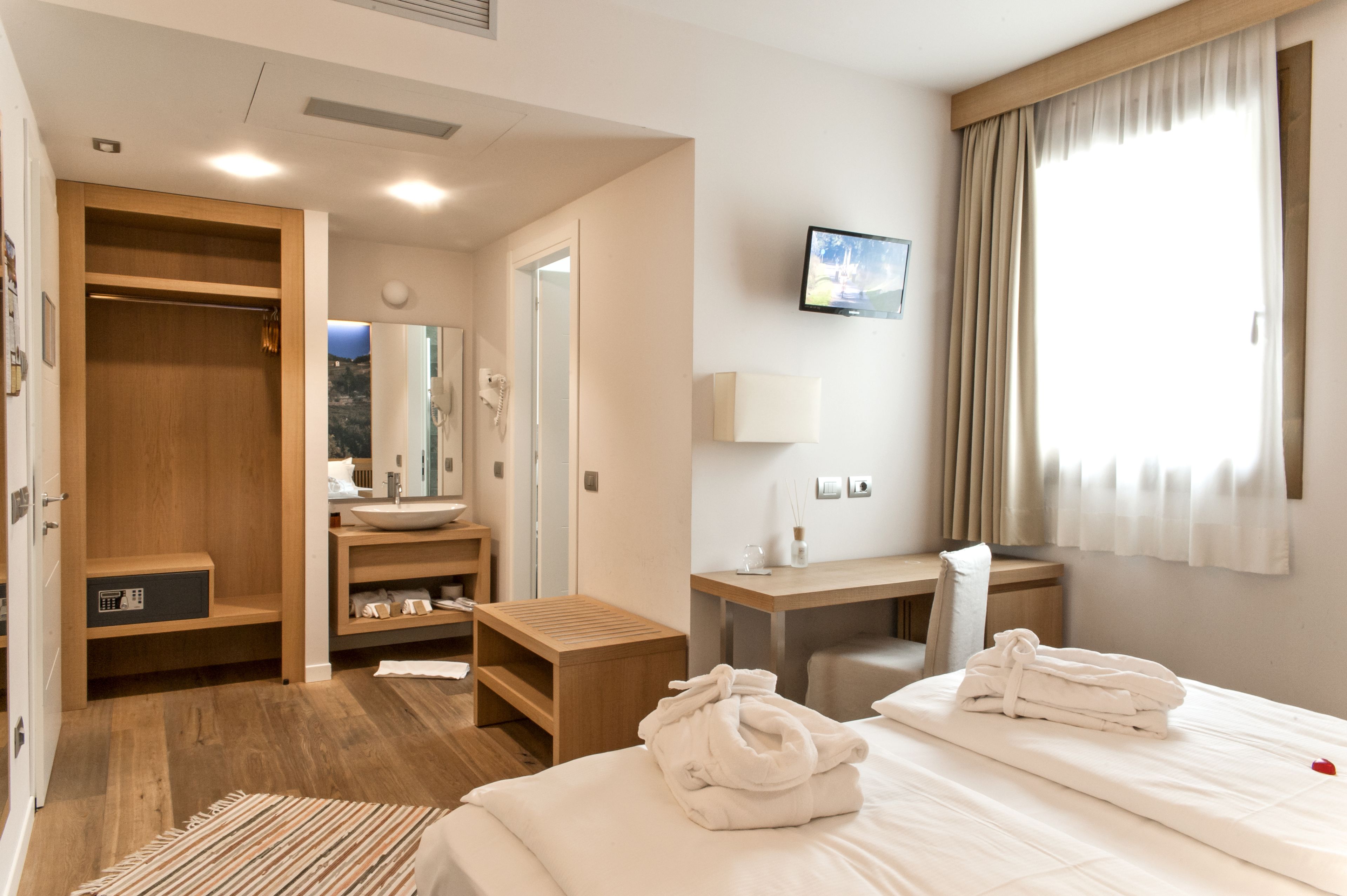 Ein Hotelzimmer mit Bett, Schreibtisch, Schminktisch mit Spiegel, Kleiderschrank und Fernseher an der Wand.