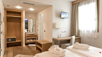 Ein Hotelzimmer mit Bett, Schreibtisch, Schminktisch mit Spiegel, Kleiderschrank und Fernseher an der Wand.
