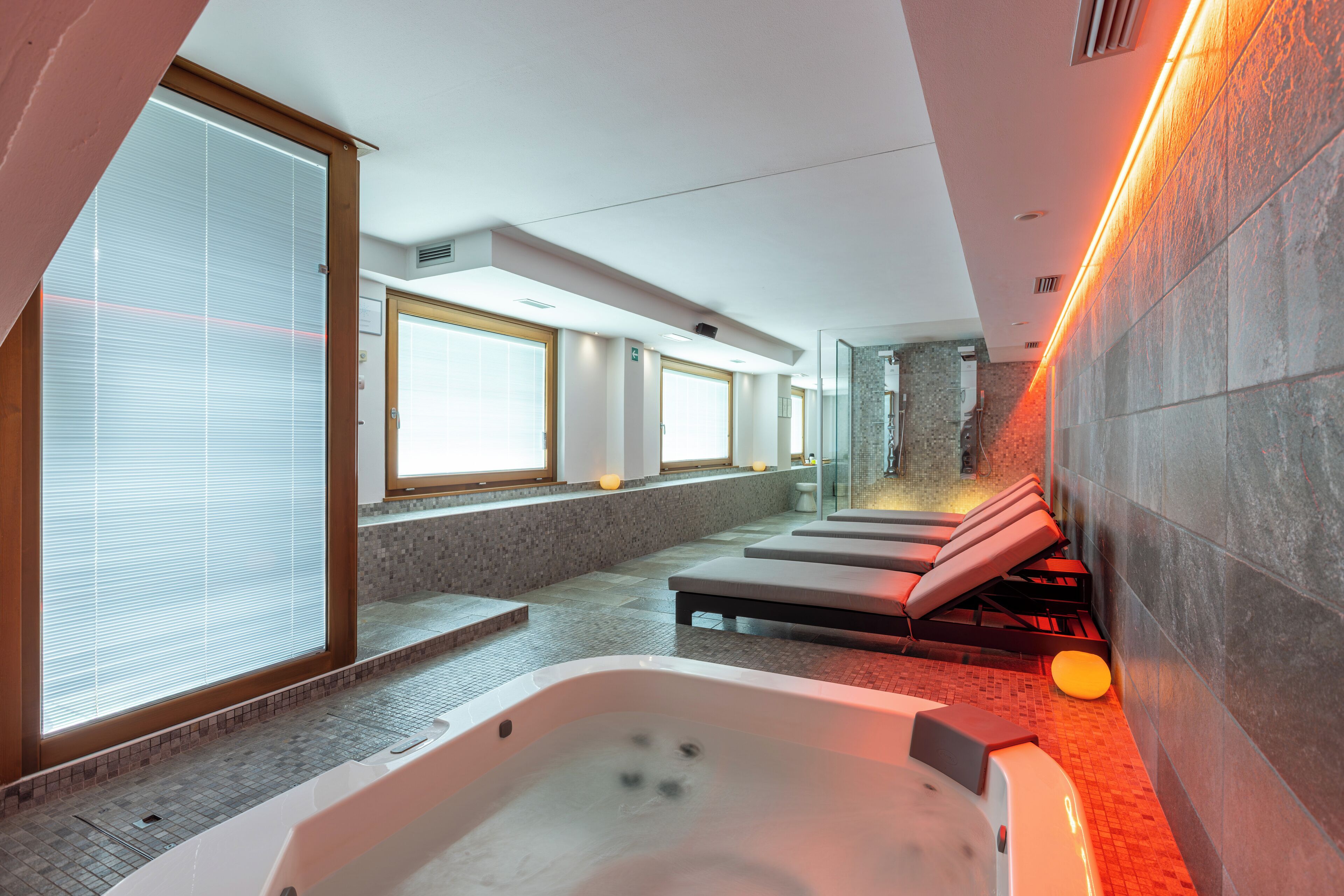 A modern spa with a large hot tub, sunlit windows, and a stone wall.