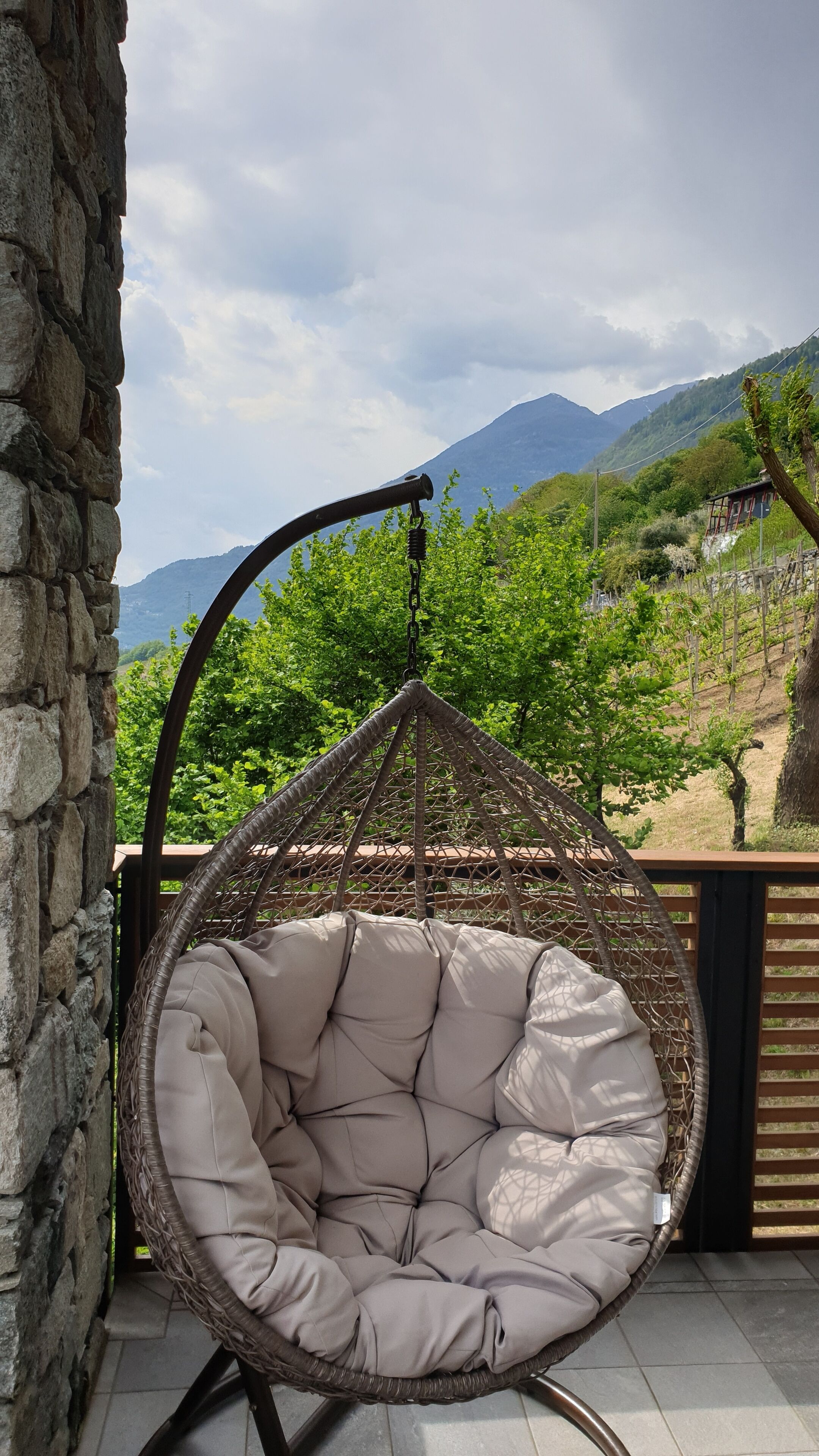 A wicker hanging chair with a cushioned seat and back, positioned on a patio with a stone wall to the left and a view of green hills and a mountain in the background.