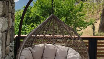 A wicker hanging chair with a cushioned seat and back, positioned on a patio with a stone wall to the left and a view of green hills and a mountain in the background.