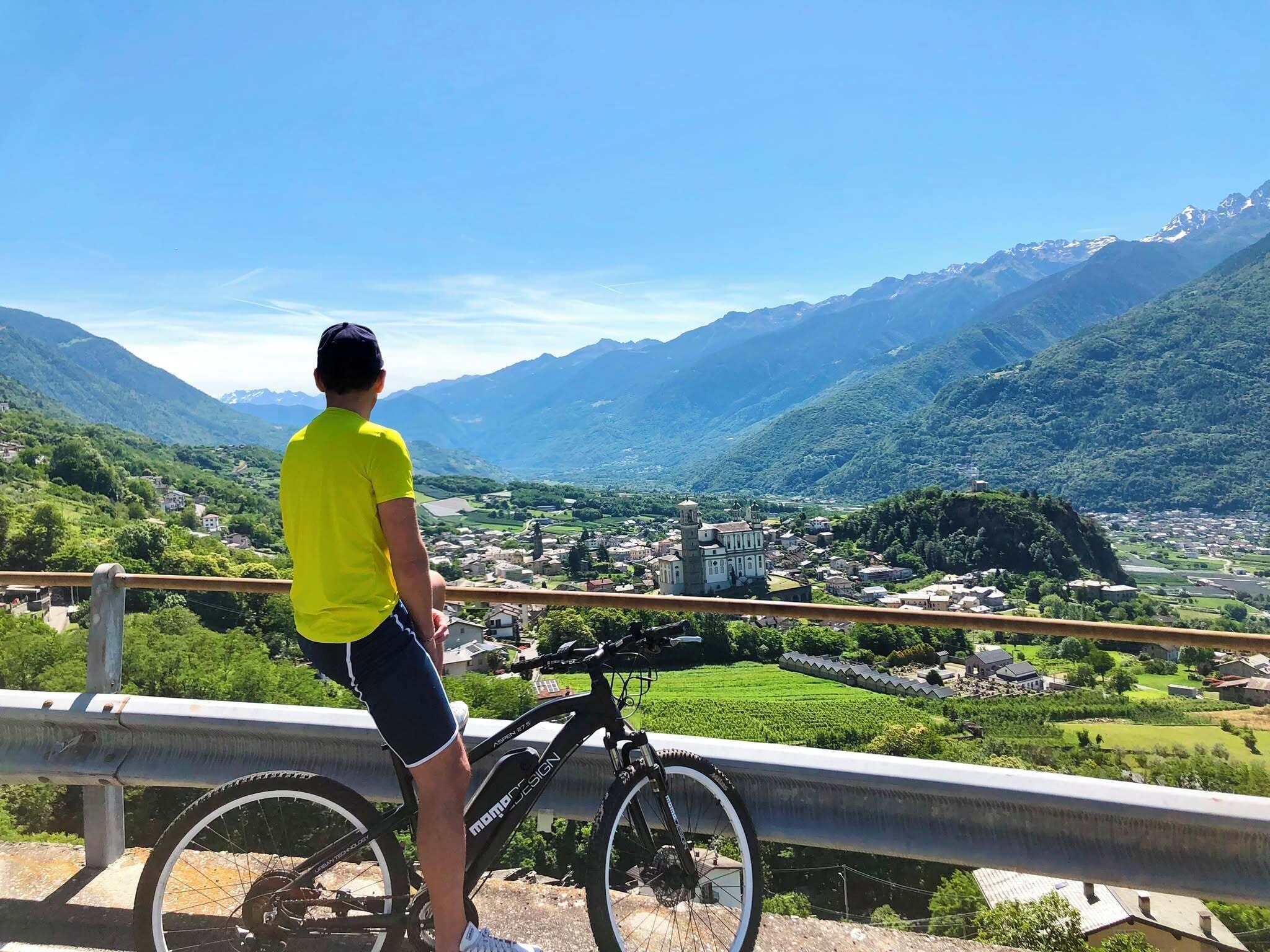 En person står bredvid en cykel och blickar ut över ett bergigt landskap med en stad i dalen.